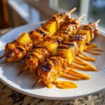 Close-up of grilled chipotle pineapple bang bang chicken skewers drizzled with spicy orange sauce on a white plate.
