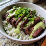 A close-up of cilantro lime steak bowls featuring sliced steak over white rice, drizzled with a creamy green sauce and topped with chopped scallions.