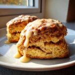 Two fluffy cinnamon bun scones topped with thick, dripping vanilla glaze sitting on a light plate.