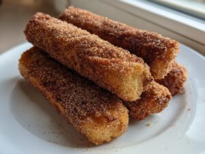 A stack of golden brown cinnamon french toast roll ups heavily coated in cinnamon sugar, resting on a white plate.