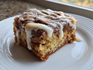 A square slice of moist cinnamon roll cake topped with thick white icing drizzle.