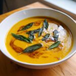 A vibrant bowl of creamy orange acorn squash soup topped with crispy fried sage leaves and brown butter swirls.