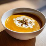A close-up of a vibrant orange autumn squash soup in a white bowl, swirled with cream and topped with toasted pumpkin seeds.