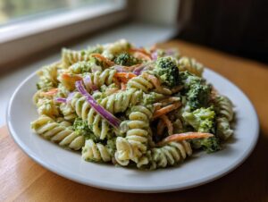 A close-up of creamy california pasta salad featuring rotini pasta, broccoli florets, shredded carrots, and red onion slices on a white plate.