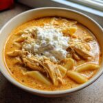 A close-up of a bowl of creamy chicken lasagna soup topped with shredded chicken, pasta, and a dollop of ricotta cheese.