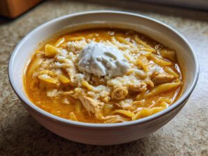 A close-up of a white bowl filled with creamy chicken lasagna soup, topped with cheese and a dollop of sour cream.