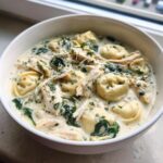 A close-up of a white bowl filled with creamy chicken tortellini soup, featuring shredded chicken, spinach, and herbs.