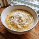 A close-up of a white bowl filled with creamy spaghetti squash soup, topped generously with shredded Parmesan cheese and black pepper.