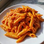 Close-up of penne pasta tossed generously in a rich, orange-hued creamy tomato pasta sauce on a white plate.