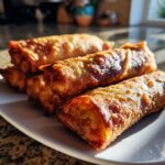 Close-up of several golden brown, crispy bacon cheeseburger egg rolls stacked on a white plate.