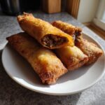 Close-up of several golden brown, crispy cheeseburger egg rolls stacked on a white plate, one is cut open showing the savory filling.