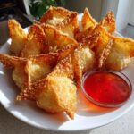 A plate piled high with golden brown, crispy crab rangoons served with a side of bright red sweet chili dipping sauce.