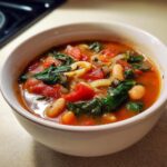 Close-up of a white bowl filled with rich, tomato-based crockpot minestrone soup featuring white beans, spinach, and pasta.