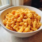 A close-up view of a white bowl filled with rich, orange extra creamy classic mac and cheese, lightly seasoned with pepper.