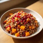 A close-up of a bowl filled with festive farro salad featuring grains, roasted orange squash cubes, and bright red pomegranate seeds.