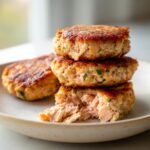 A stack of three golden-brown salmon patties, with the bottom one broken open to show the flaky, pink interior of the salmon patties.