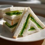 A stack of crustless, triangular cucumber sandwiches filled with thin cucumber slices and cream cheese.