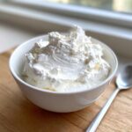 A white bowl filled with fluffy, homemade whipped cream, perfect for an easy fruit dip, resting on a wooden board.