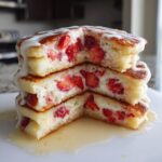 A stack of three fluffy strawberry cheesecake pancakes cut in half, showing the creamy filling and strawberries inside, drizzled with glaze.