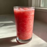 A tall glass filled with bright red, icy frozen strawberry lemonade, showing condensation on the outside.