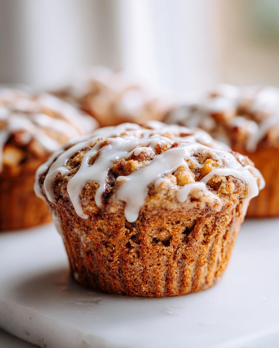 A close-up of a perfectly baked apple fritter muffin topped with a thick white glaze drizzle.
