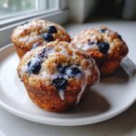 Three freshly baked lemon blueberry cottage cheese muffins topped with a sweet white glaze, sitting on a white plate.