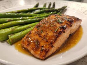 A perfectly cooked, glazed salmon fillet served alongside bright green roasted asparagus on a white plate.