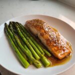 A perfectly cooked salmon fillet topped with a shiny glaze, served next to bright green asparagus spears on a white plate.