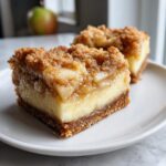 Two decadent gluten free apple cheesecake bars with a thick layer of creamy filling and crumb topping on a white plate.