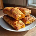 Four crispy, golden brown cheeseburger egg rolls stacked on a white plate, showing melted cheese filling.