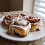 Four freshly baked, gooey cinnamon rolls covered in thick white icing sitting on a white plate.