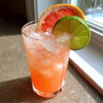 A tall glass filled with a bubbly, pink grapefruit lime spritzer, ice, and garnished with a slice of grapefruit and lime.
