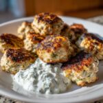 Close-up of golden brown greek chicken meatballs served with a dollop of creamy tzatziki sauce on a white plate.