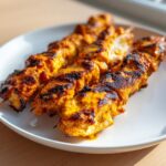 Close-up of three juicy, grilled bang bang chicken skewers with char marks resting on a white plate.