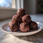 A pyramid stack of rich, dark high protein coffee truffles coated in finely chopped nuts or seeds, sitting on a white plate.