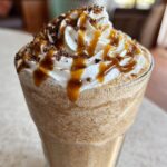 Close-up of a tall glass filled with a blended homemade caramel frappe, topped with whipped cream, caramel drizzle, and chocolate shavings.