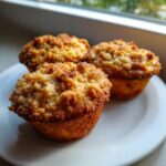 Three golden brown homemade peach cobbler muffins topped with a thick, crunchy streusel topping, resting on a white plate.
