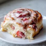 A close-up of a single, gooey strawberry cinnamon roll slice topped with white icing and visible strawberry pieces.