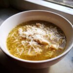 A close-up of a white bowl filled with hot italian penicillin soup, featuring shredded chicken and topped generously with grated Parmesan cheese.