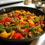 Close-up of Italian sausage and peppers (red, yellow, and green) sizzling together in a dark cast-iron skillet.