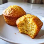 Two golden keto lemon muffins on a white plate, one with a bite taken out showing the moist interior texture.
