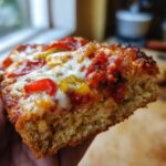 A hand holding a thick slice of keto pizza recipe, showing the fluffy crust, melted cheese, pepperoni, and peppers.
