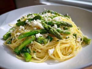 Close-up of a bright lemon capellini salad tossed with vibrant green asparagus spears and crumbled white feta cheese.