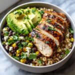 A close-up of a lemon chili grilled chicken bowls featuring sliced chicken breast over rice, black beans, corn, avocado, and cheese.