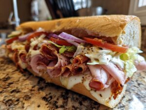 Close-up of a fully loaded italian grinder sub featuring layers of deli meats, melted cheese, lettuce, tomato, and red onion.