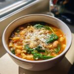 Close-up of a steaming bowl of minestrone soup topped with fresh spinach, white beans, pasta, and grated Parmesan cheese.