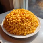 A large, perfectly domed serving of classic creamy mac and cheese, coated in bright orange cheese sauce, sits on a white plate.