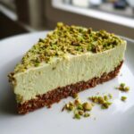 A perfect slice of no bake pistachio cheesecake with a thick, light green filling and a dark crust, topped generously with chopped pistachios.