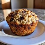 A single, perfectly baked oatmeal chocolate chip morning muffin with a golden-brown, textured top, resting on a white plate.