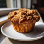 A close-up of a golden brown peach cobbler muffins with a thick, sugary streusel topping, sitting on a white plate.